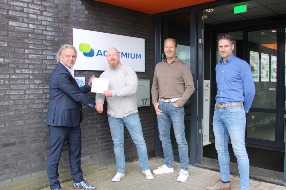 Medewerkers van het Graafschap College en Actemium staan voor het Actemium-kantoor in Doetinchem tijdens de overhandiging van een gevelschild. Op de foto schudden twee personen elkaar de hand terwijl een bord van het Graafschap College wordt overhandigd. Op de achtergrond zijn het Actemium-logo en de entree van het pand zichtbaar.