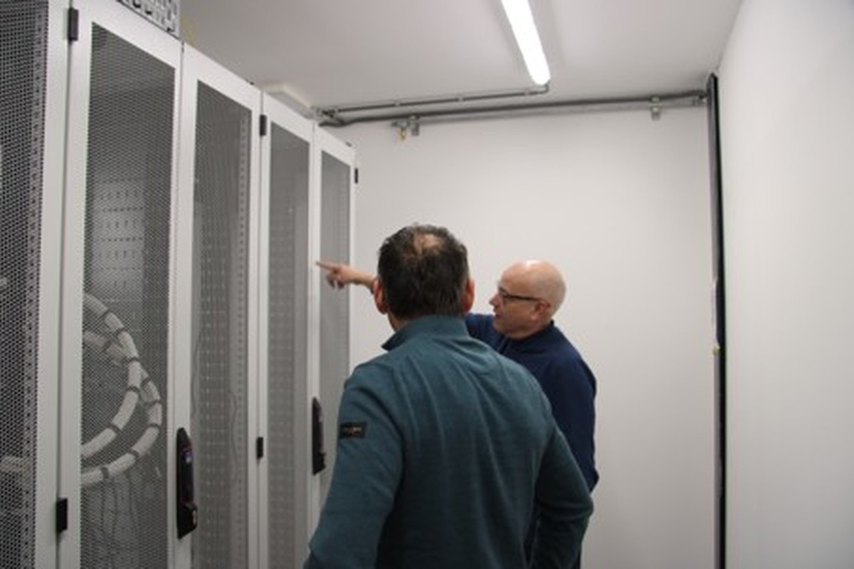 Twee medewerkers inspecteren serverracks en bekabeling in een afgesloten technische ruimte voor communicatie- en datanetwerken.