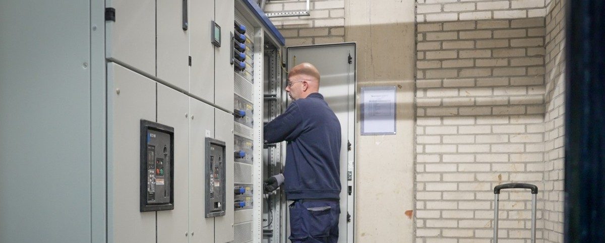 Monteur werkt aan een geopende elektrische verdeelkast in een technische ruimte, met gereedschapskoffer op de vloer.