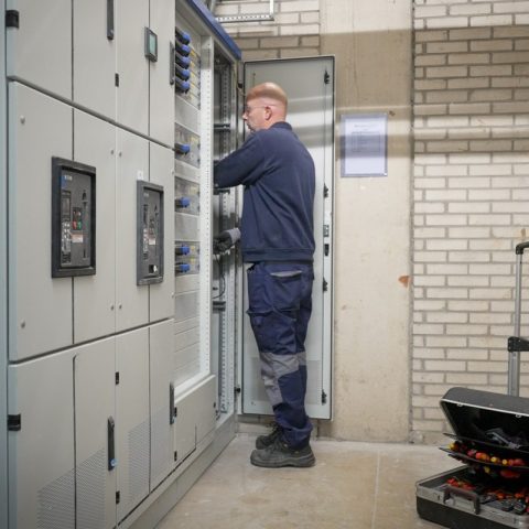 Monteur werkt aan een geopende elektrische verdeelkast in een technische ruimte, met gereedschapskoffer op de vloer.