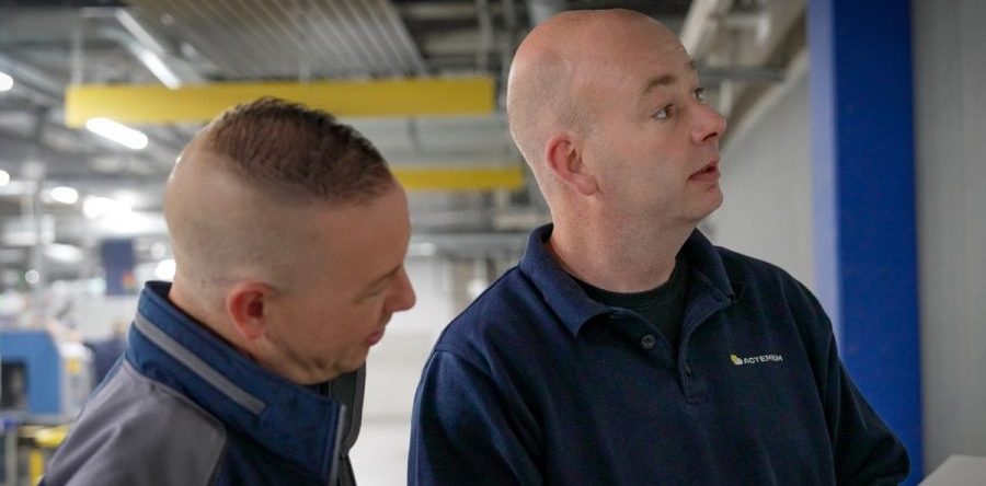 Twee medewerkers controleren technische documenten en installaties in een industriële omgeving met machines en leidingwerk op de achtergrond.