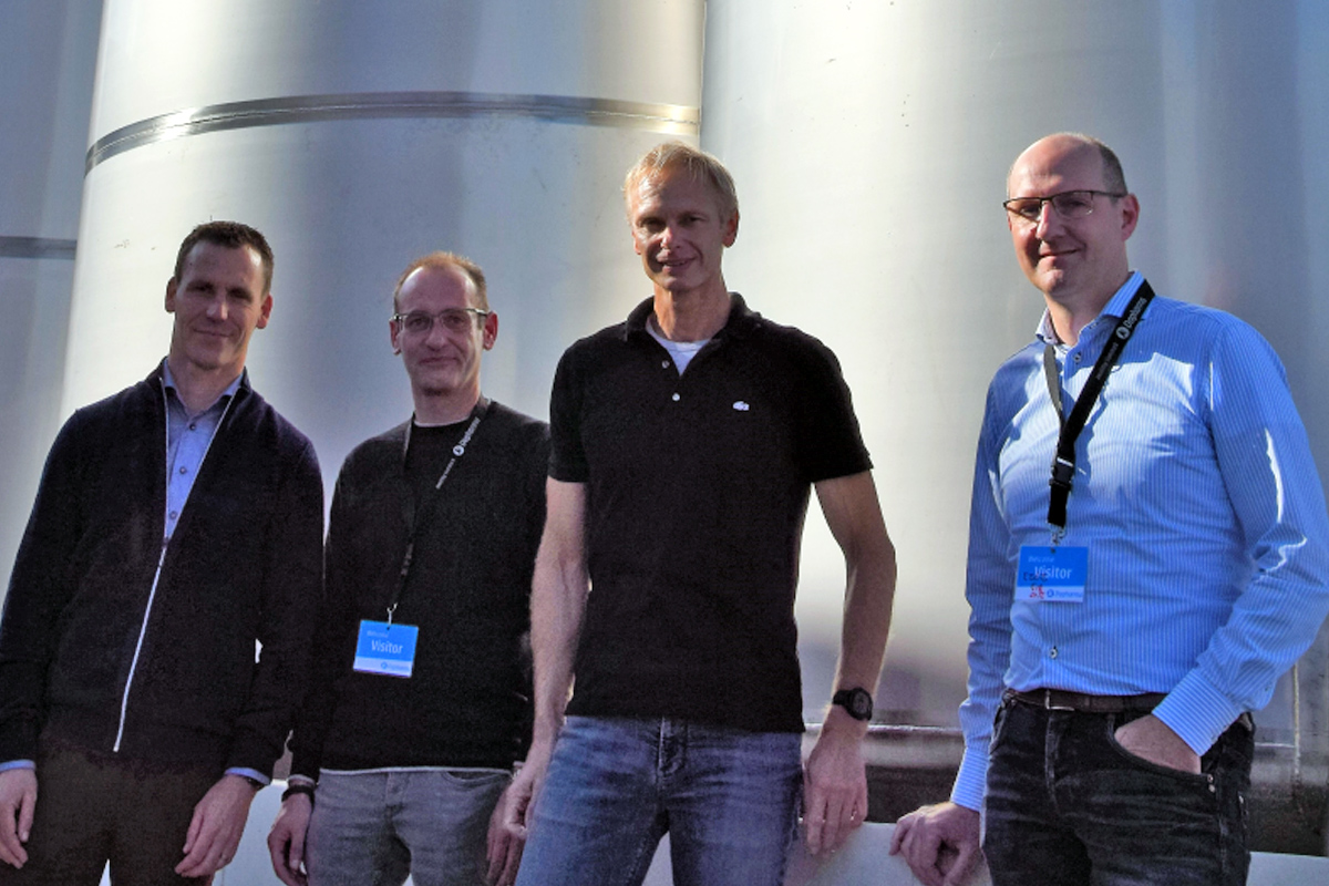 Vier mannen poseren naast elkaar voor grote, glanzende metalen industriële tanks. Van links naar rechts staan Marc Niessen (Life Sciences), Eugene Akkermans (Dopharma) met een blauw 'Visitor' keycord, Ron de Bruijn (Actemium) in een zwart poloshirt en spijkerbroek, en Mario Willaert (Siemens) in een blauw gestreept overhemd met eveneens een blauw bezoekerspasje.