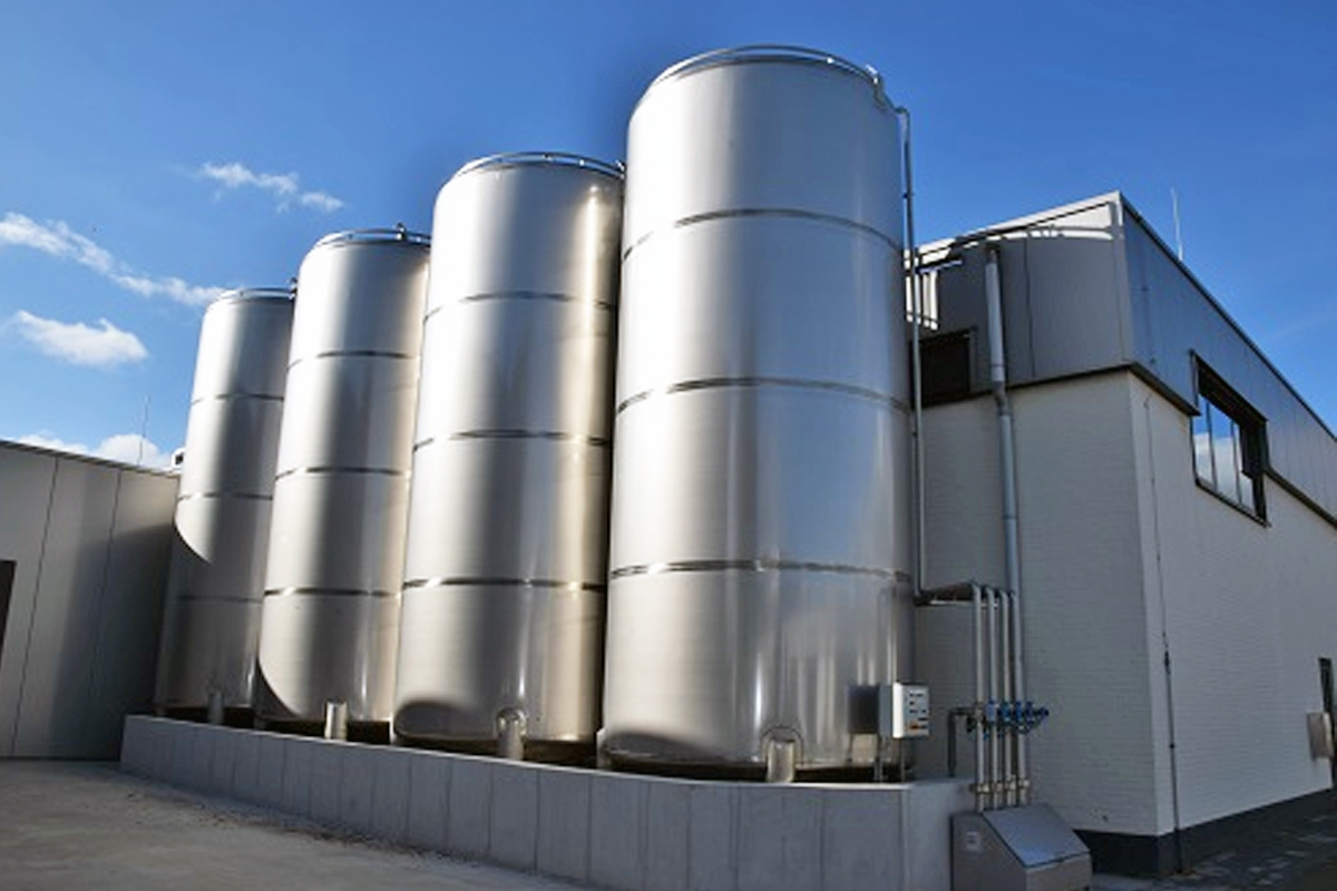 Vier grote, glanzende roestvrijstalen opslagtanks van Dopharma staan opgesteld in een betonnen opvangbak naast een modern fabrieksgebouw onder een strakke blauwe lucht.