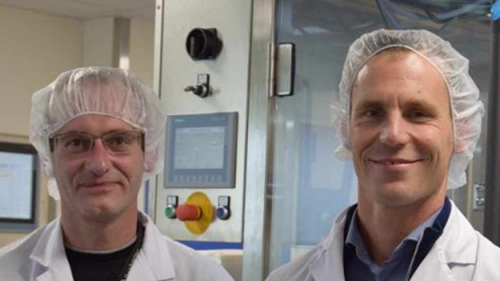 Eugene Akkermans van Dopharma en Marc Niessen van Life Sciences dragen witte laboratoriumjassen en haarnetjes voor een industrieel bedieningspaneel in de fabriek.