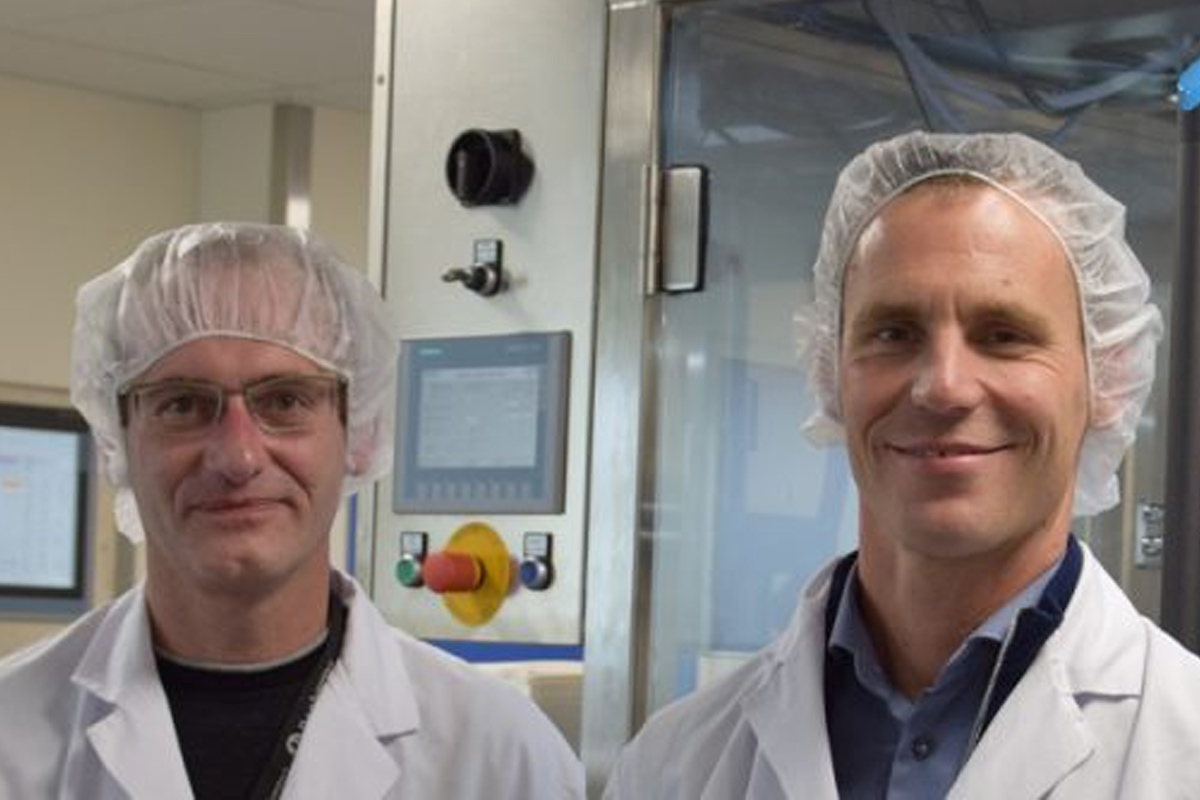 Eugene Akkermans van Dopharma en Marc Niessen van Life Sciences dragen witte laboratoriumjassen en haarnetjes voor een industrieel bedieningspaneel in de fabriek.