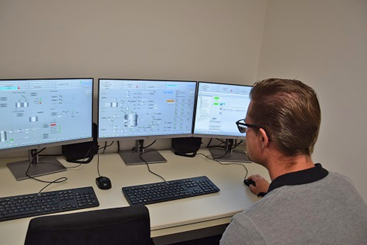 Een operator van Actemium zit achter een bureau en controleert het productieproces van Dopharma via drie computerschermen met procesautomatiseringsdiagrammen.