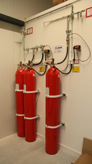 Actemium realiseert beveiligingsinstallaties voor een veilige gebouwomgeving. Op de afbeelding is een noodverlichtingsarmatuur met vluchtroutepictogram te zien, gemonteerd aan een wand met zichtbare bekabeling en leidingwerk.
