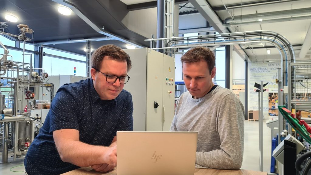 Twee medewerkers analyseren procesdata op een laptop in een industriële productieomgeving met installaties, leidingen en automatiseringsapparatuur. Actemium in Nederland.