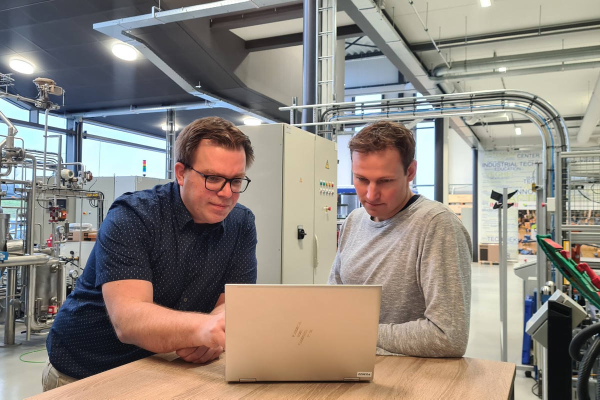 Twee medewerkers analyseren procesdata op een laptop in een industriële productieomgeving met installaties, leidingen en automatiseringsapparatuur. Actemium in Nederland.