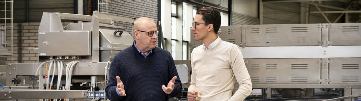 Twee medewerkers bespreken een industriële installatie naast een geautomatiseerde productielijn met transportsystemen en RVS-machines in een productieomgeving.