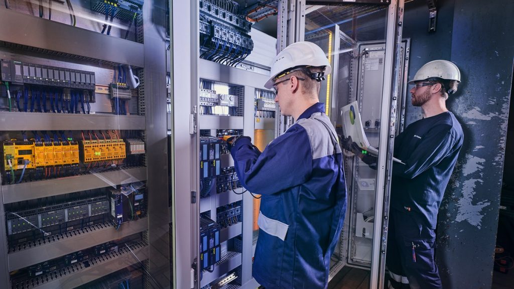 Actemium monteur werkt met een laptop in een technische ruimte naast een geopende schakelkast met bekabeling en industriële installaties.