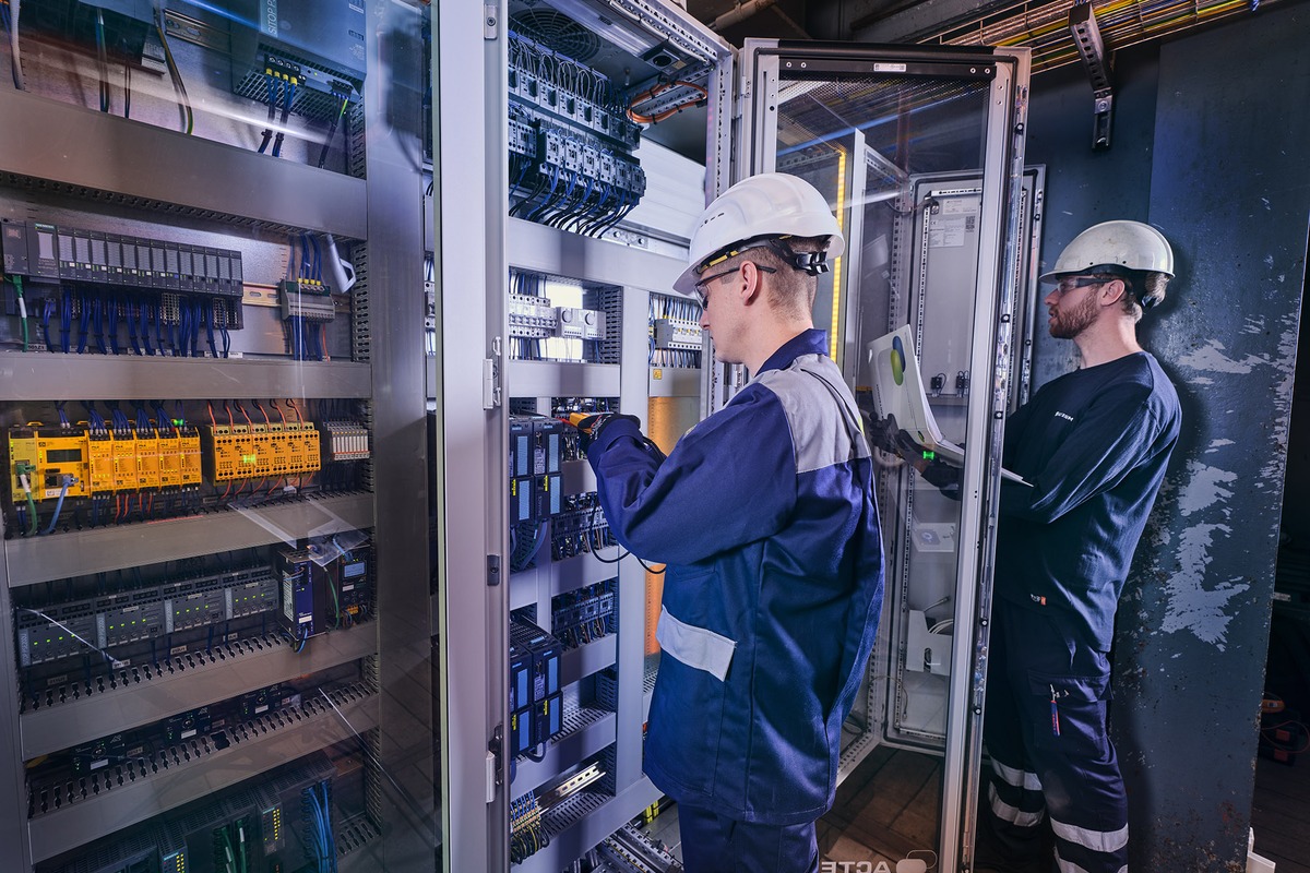 Actemium monteur werkt met een laptop in een technische ruimte naast een geopende schakelkast met bekabeling en industriële installaties.