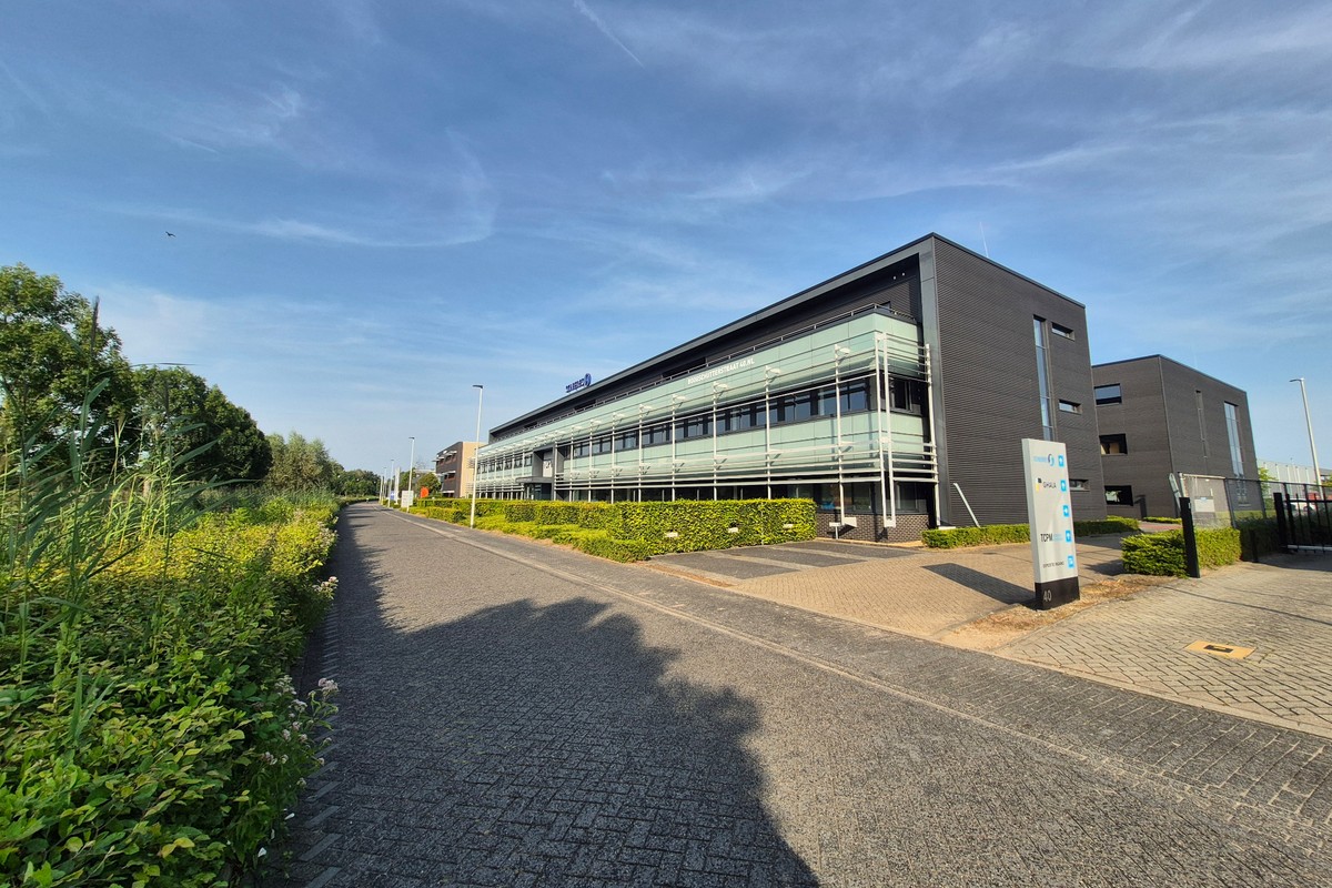 Modern pand van Actemium in Apeldoorn met glazen gevel en omliggend groen langs een toegangsweg.