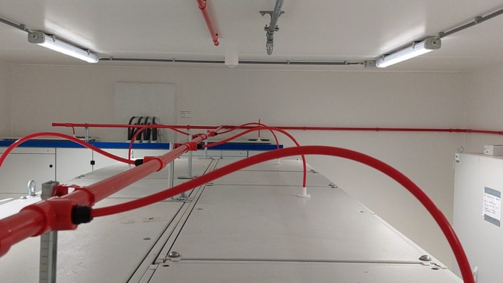 Brandmeldinstallatie in een technische ruimte met rode detectieleidingen boven paneelkasten. Actemium Building Technologies realiseert en onderhoudt brandmeldinstallaties die bijdragen aan een veilige bedrijfsomgeving.